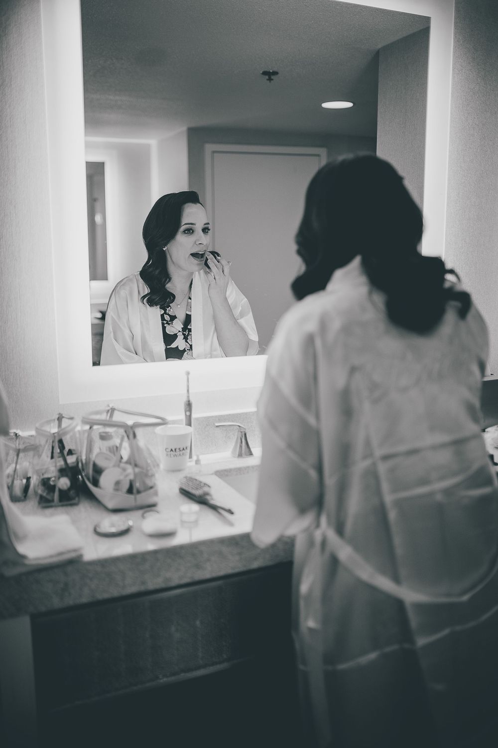 Black and white photo showing reflection in mirror with makeup and beauty products on vanity counter.