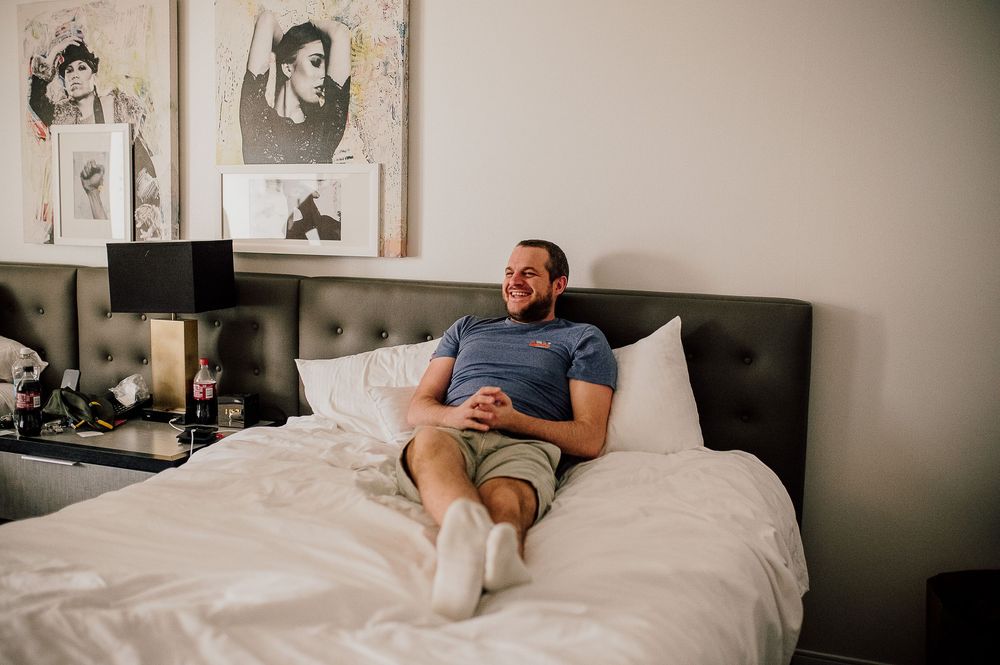Someone relaxes on a white bed in a casual t-shirt and shorts with framed photos on the wall behind them.