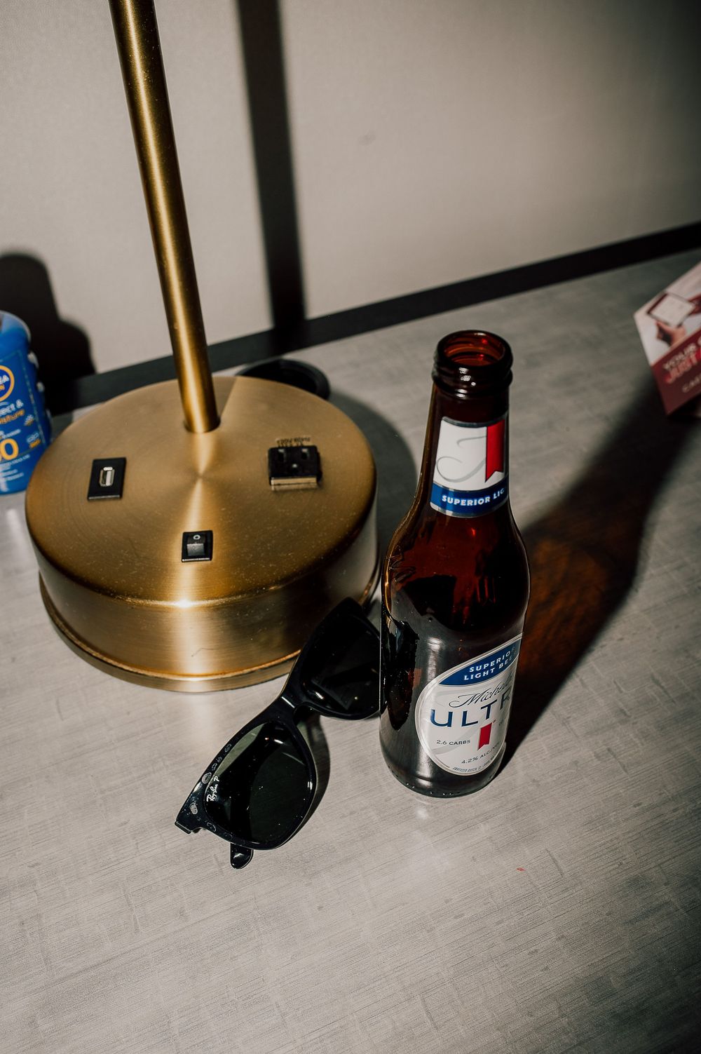 A gold desk lamp base sits beside a bottle of Michelob Ultra beer and black sunglasses on a gray surface.