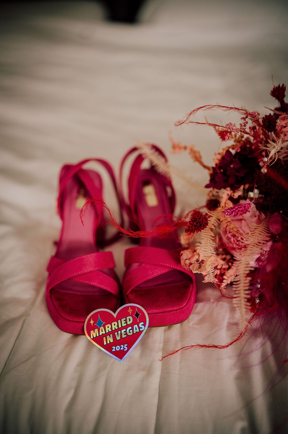 Pink wedding shoes with heart-shaped decorations and dried flowers arranged on white fabric in a romantic setting.