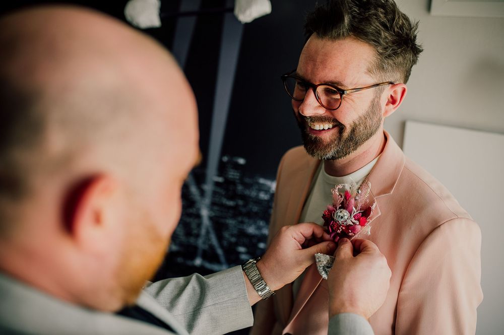 Someone helps adjust a pink floral boutonniere on a light beige suit jacket during a formal occasion.