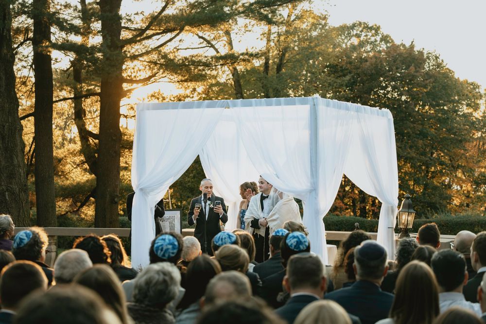 Outdoor wedding ceremony under white canopy with guests seated on lawn during golden sunset hour in wooded setting.