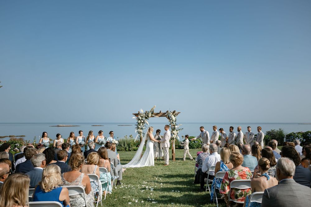 Outdoor wedding ceremony by the ocean with guests seated in white chairs and a wooden arch decorated with flowers.