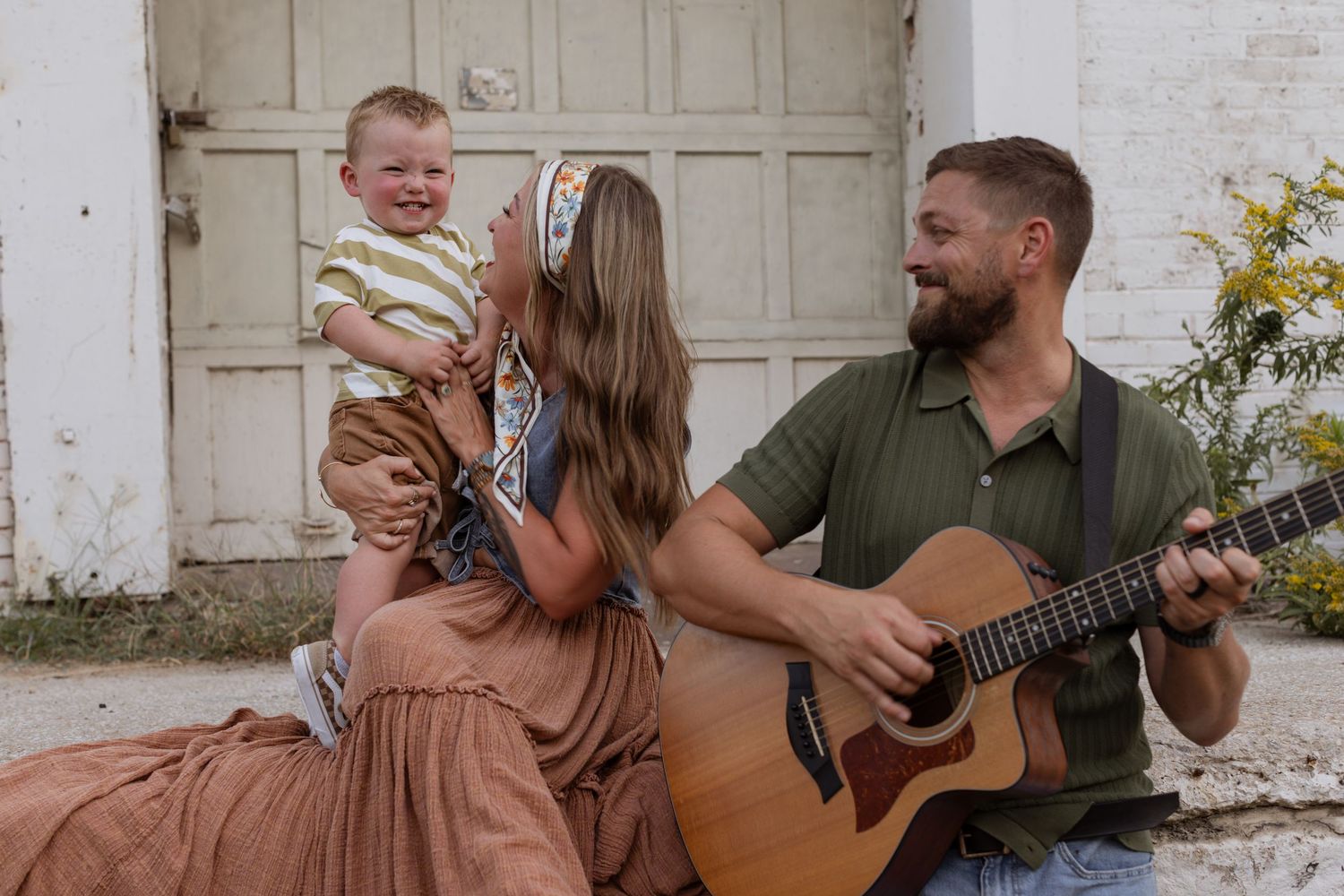 A woman tickles her toddler son and looks up at him while her husband stands next to them smiling and playing guitar as he looks on.