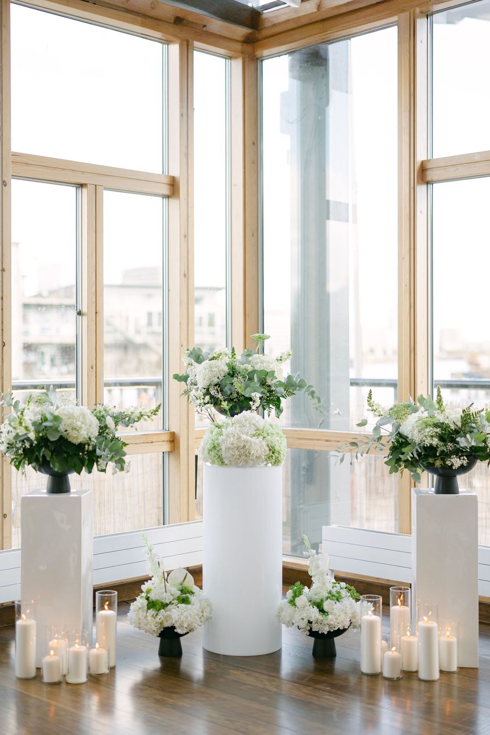 White floral arrangements in modern vases line a bright sunlit room with floor-to-ceiling windows and glowing candles.