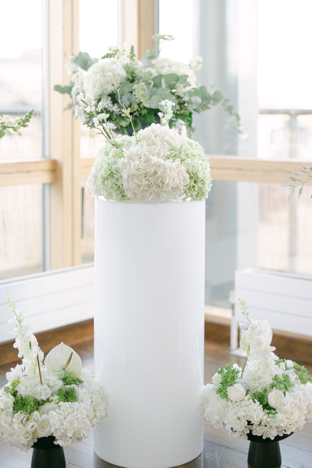White hydrangea flowers arranged in modern white cylindrical vases along a bright window sill.