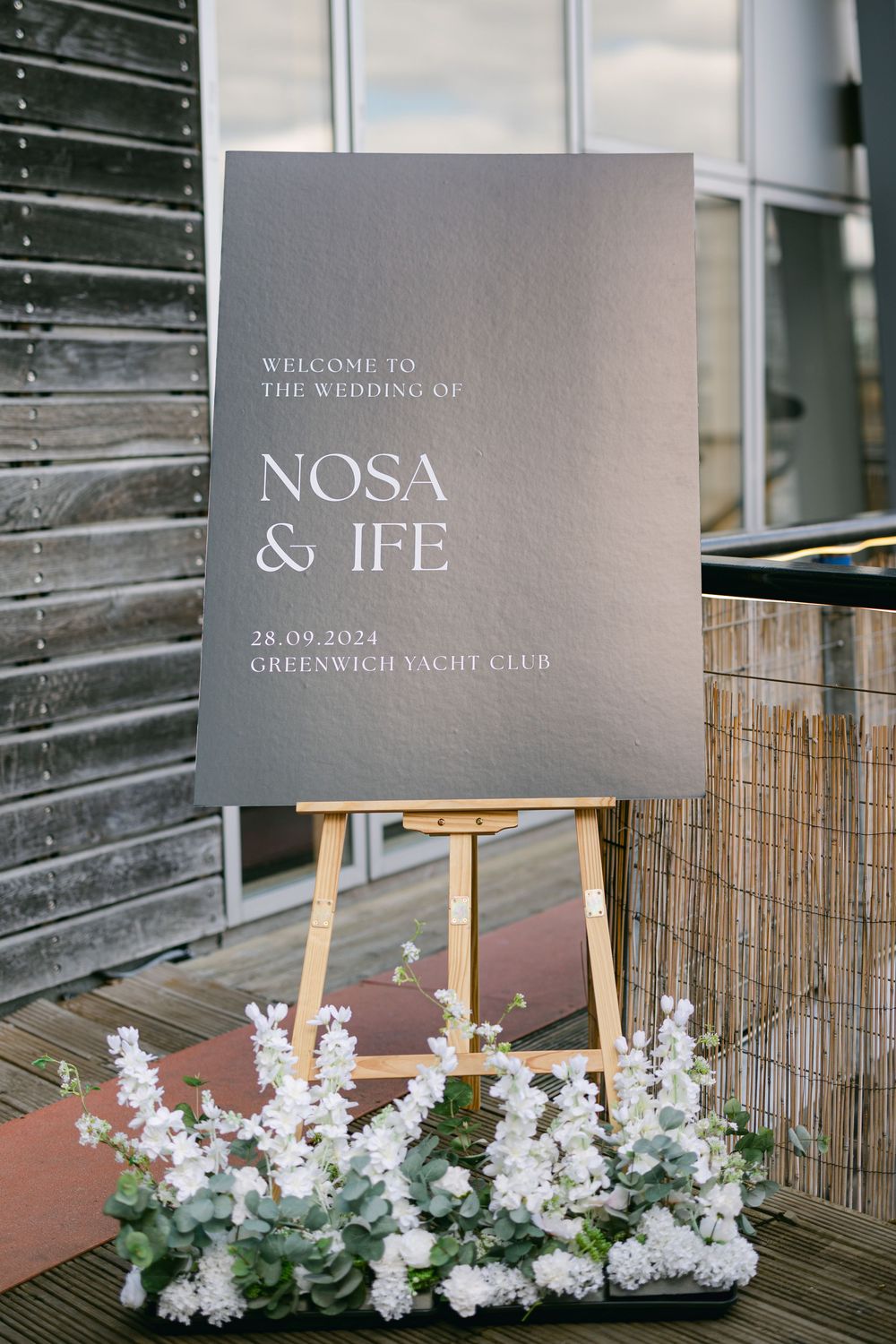 A minimalist gray wedding welcome sign for Nosa & Ife decorated with white flowers at Greenwich Yacht Club.