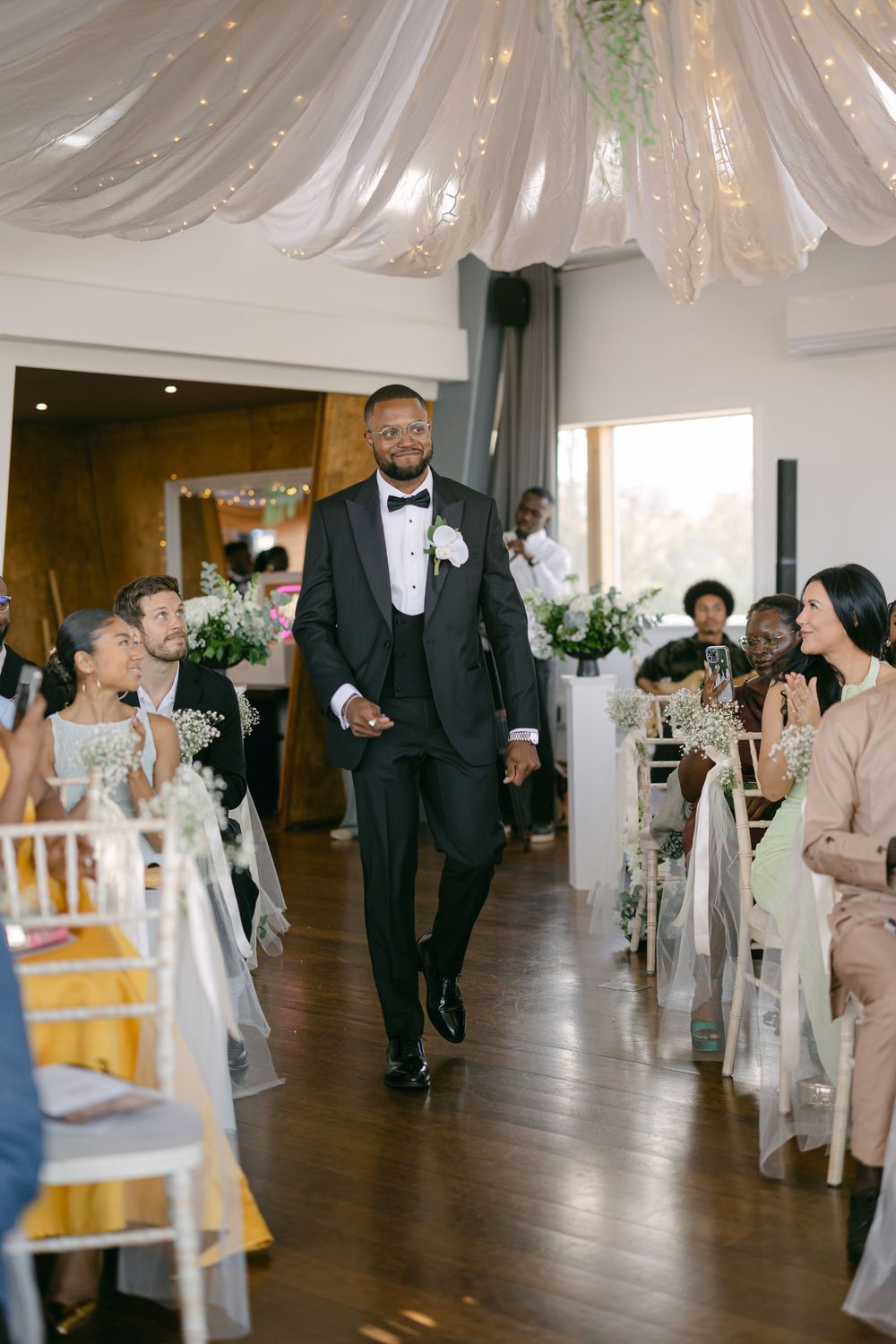 Wedding guests seated in white chairs watch as people walk down a decorated indoor aisle with draped fabric and lighting.