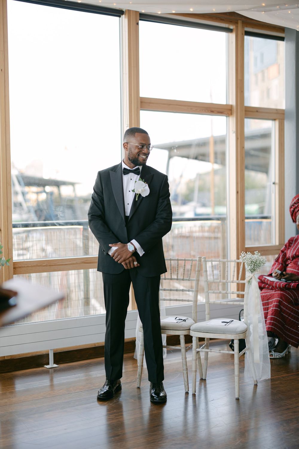 Person in formal black tuxedo stands alone in a modern venue with large windows and wooden floors.