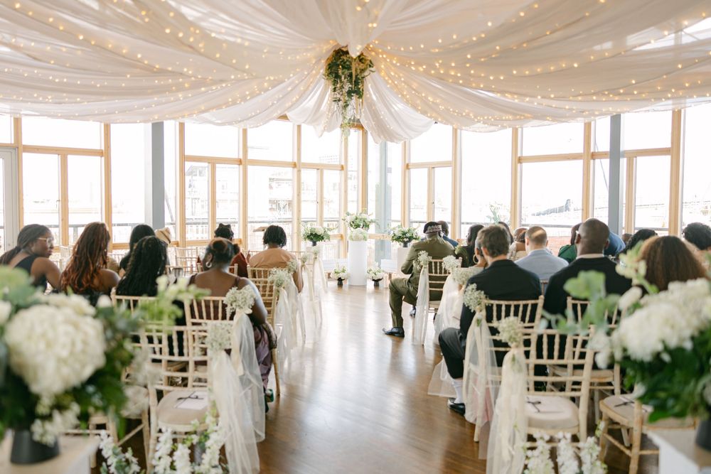 Indoor wedding ceremony setup with white chairs, floral decorations and twinkling lights in an elegant venue with large windows.