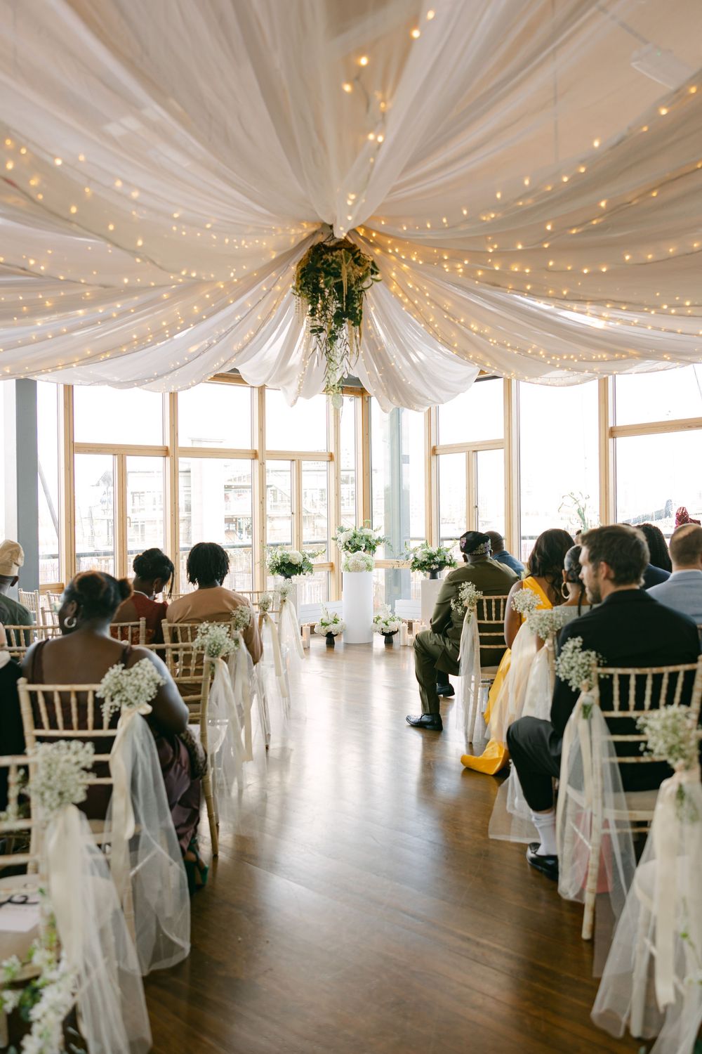 Elegant wedding venue with white draped ceiling, string lights and wooden floors set up with chiavari chairs for ceremony.