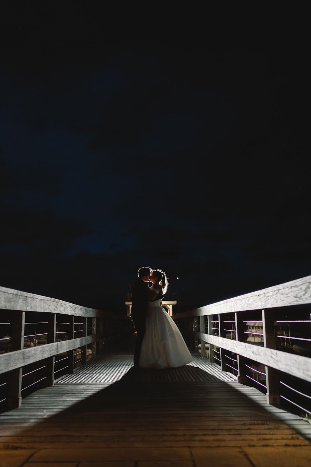 Dark wooden boardwalk lit by railing lights stretches into the night with geometric shadows and lines.