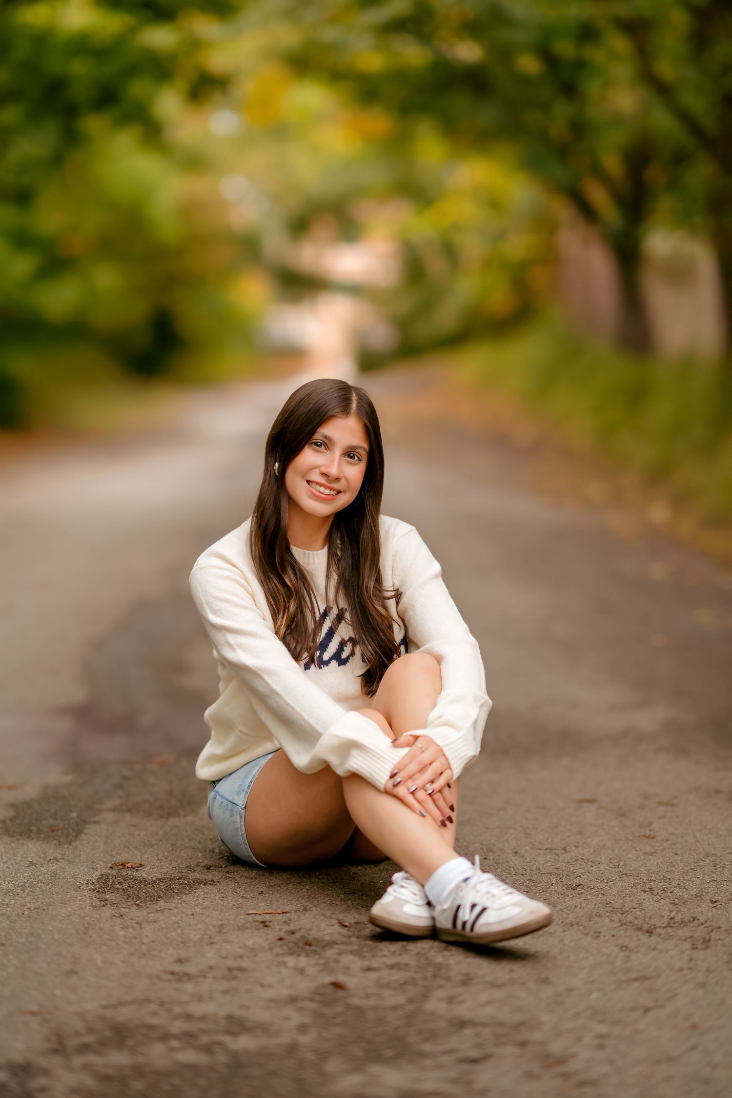 A person in a white sweater and denim shorts sits casually on a tree-lined path in an artistic photo series.