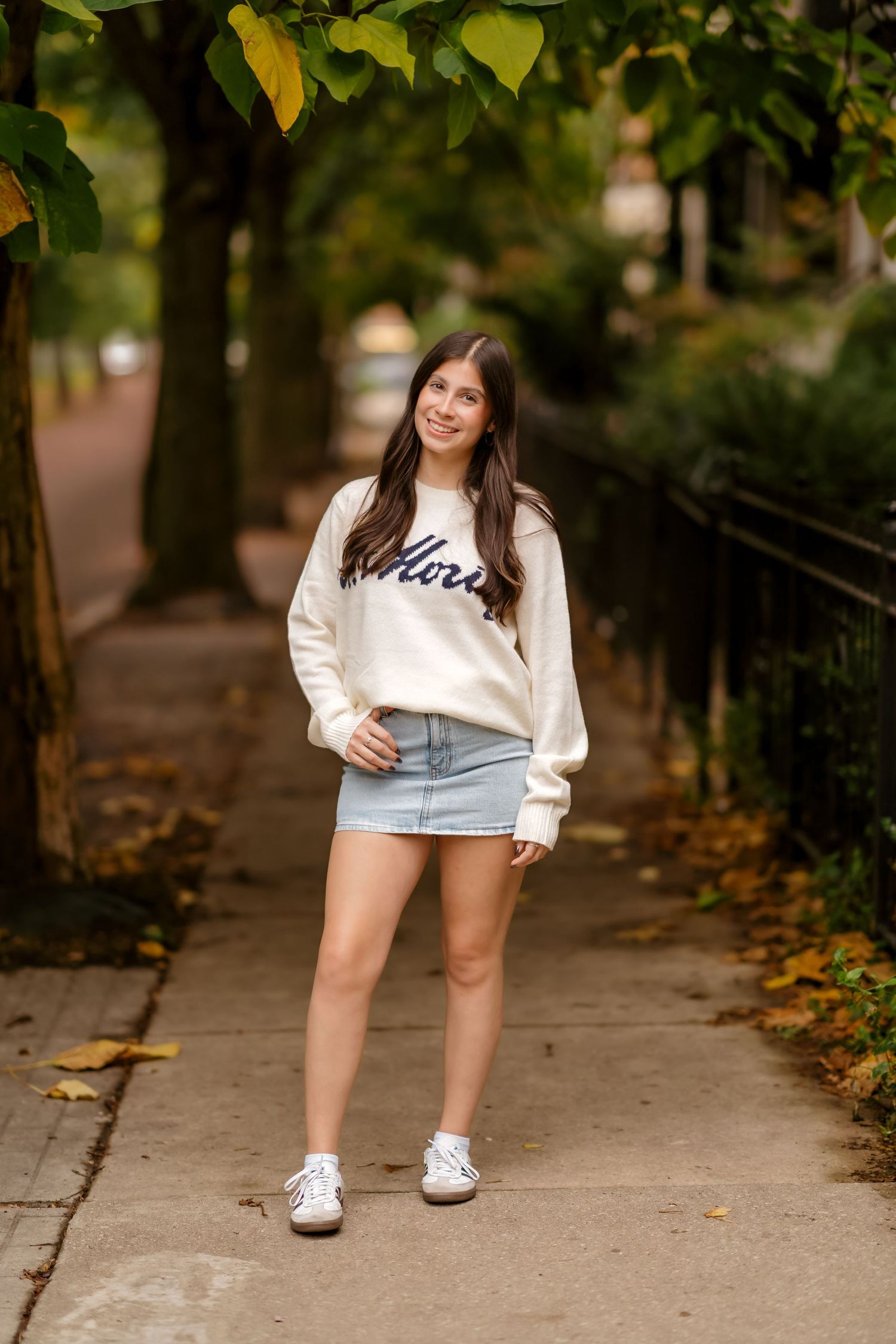 Person in a cream colored sweatshirt and denim shorts poses playfully on a sidewalk with autumn leaves.