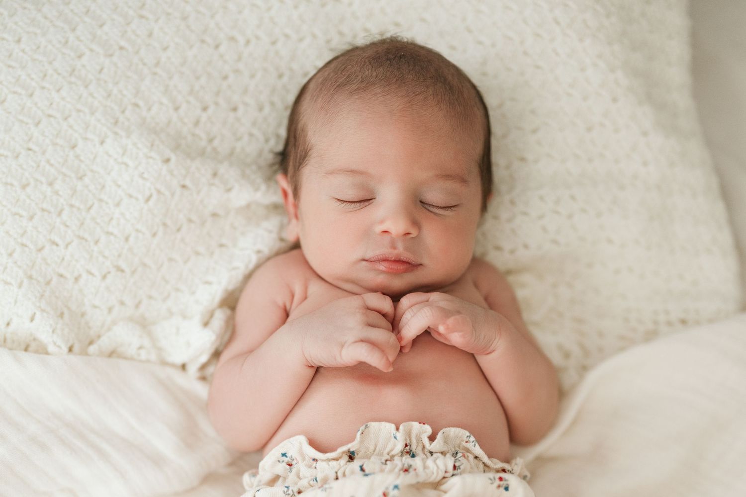 Baby in white outfit sits on blanket between hay bales in golden wheat field at sunset.