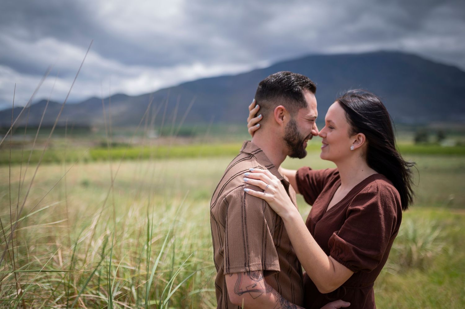 Couple sharing a romantic moment outdoors with mountains and green fields in the background under a cloudy sky.
