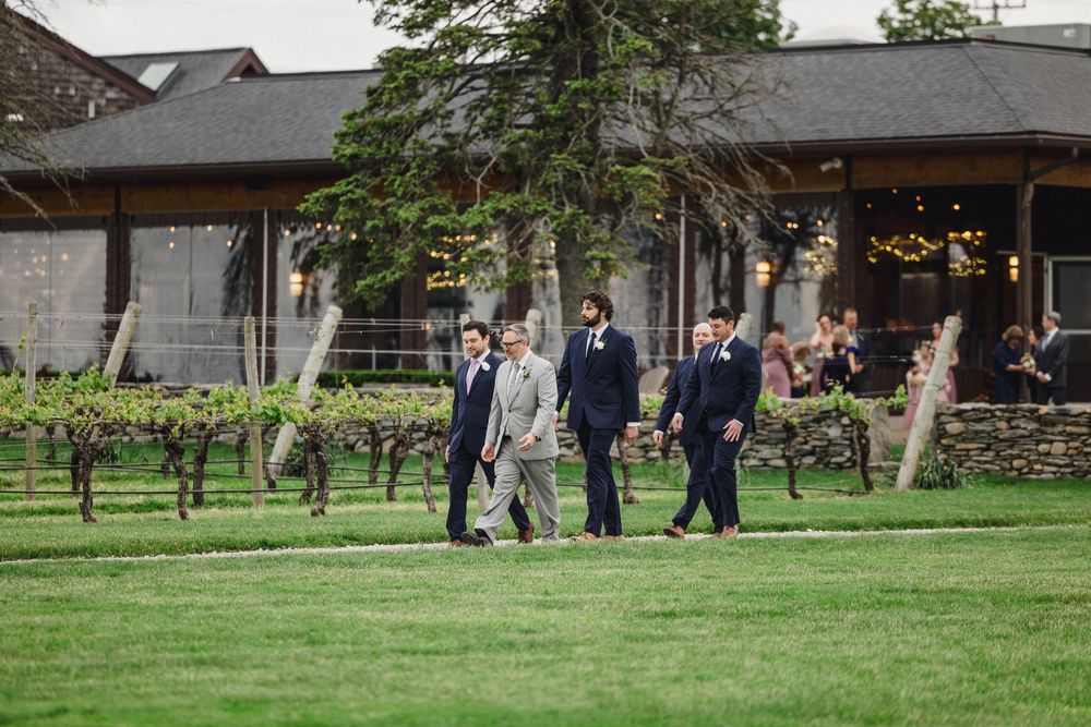 Wedding party members walk through a vineyard near a rustic building on a cloudy day.