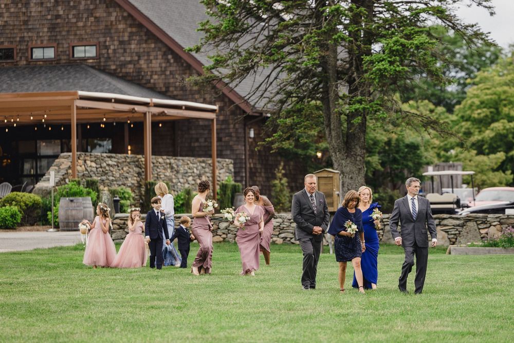 Wedding party walking along a rustic stone building with string lights and greenery decorations.