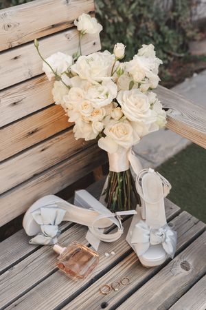 Wedding accessories including white roses, bridal shoes, and perfume arranged on wooden planks.