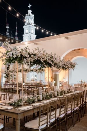 An elegant outdoor wedding reception setup with string lights and floral centerpieces under arched architecture.