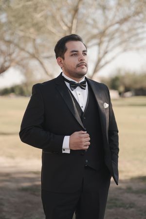 A groom in a classic black tuxedo and bow tie poses outdoors during a wedding photoshoot.