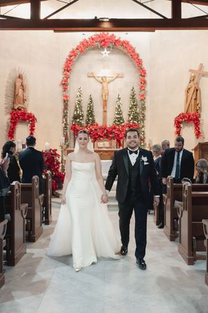 Newlyweds walk down the church aisle after their ceremony under a red floral arch.