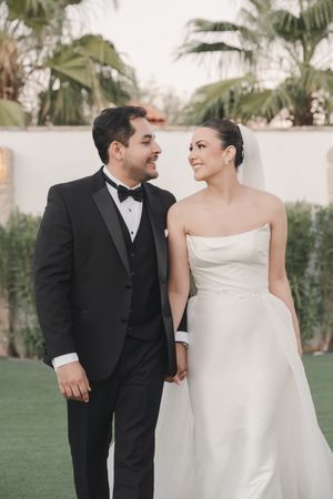 A bride in strapless gown and groom in tuxedo share a loving glance outdoors.