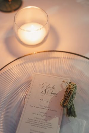 A wedding menu card with calligraphy sits on a plate next to a glowing candle.