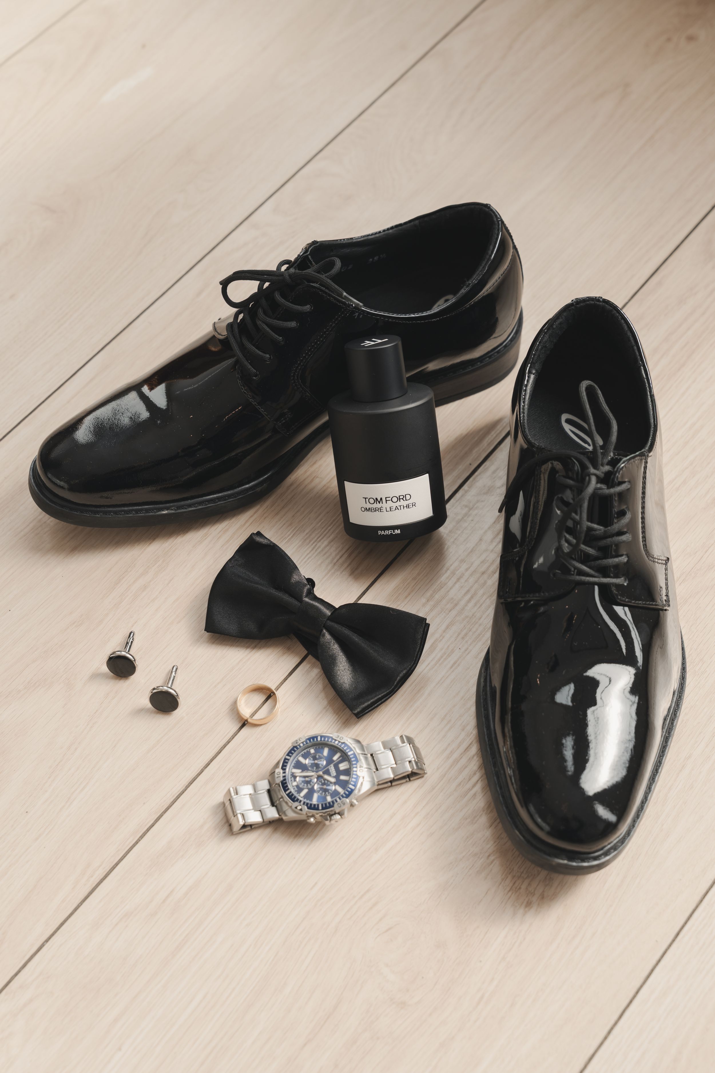 Groom's formal black dress shoes and accessories arranged on light wooden floor.
