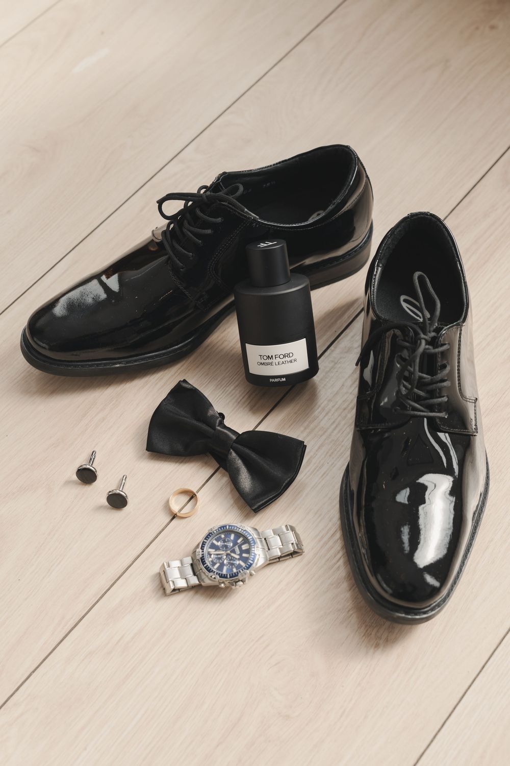 Groom's formal black dress shoes and accessories arranged on light wooden floor.
