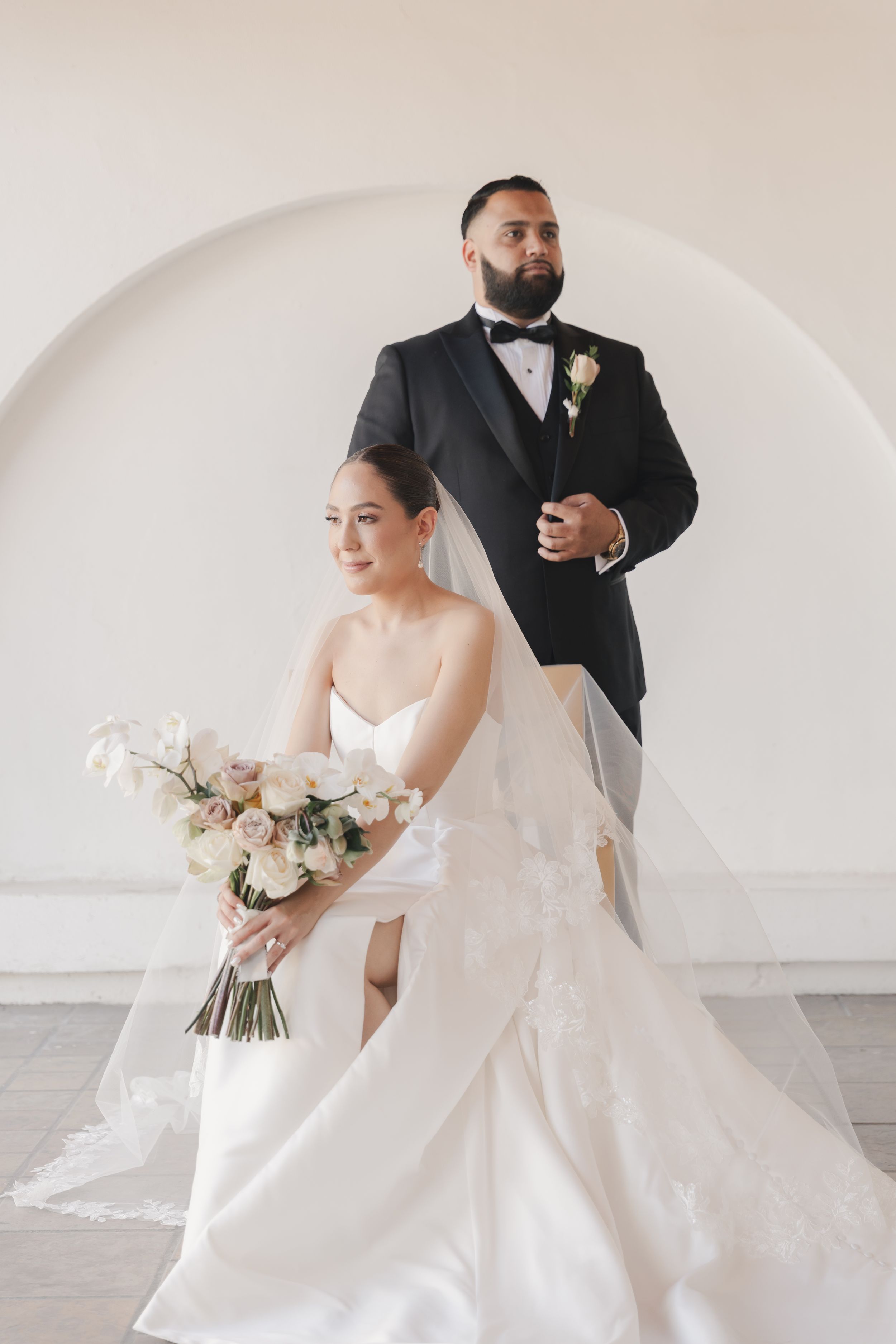 Elegant wedding couple poses under white archway in classic formal attire.