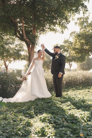 Joyful wedding couple dances in garden setting with natural sunlight filtering through trees.