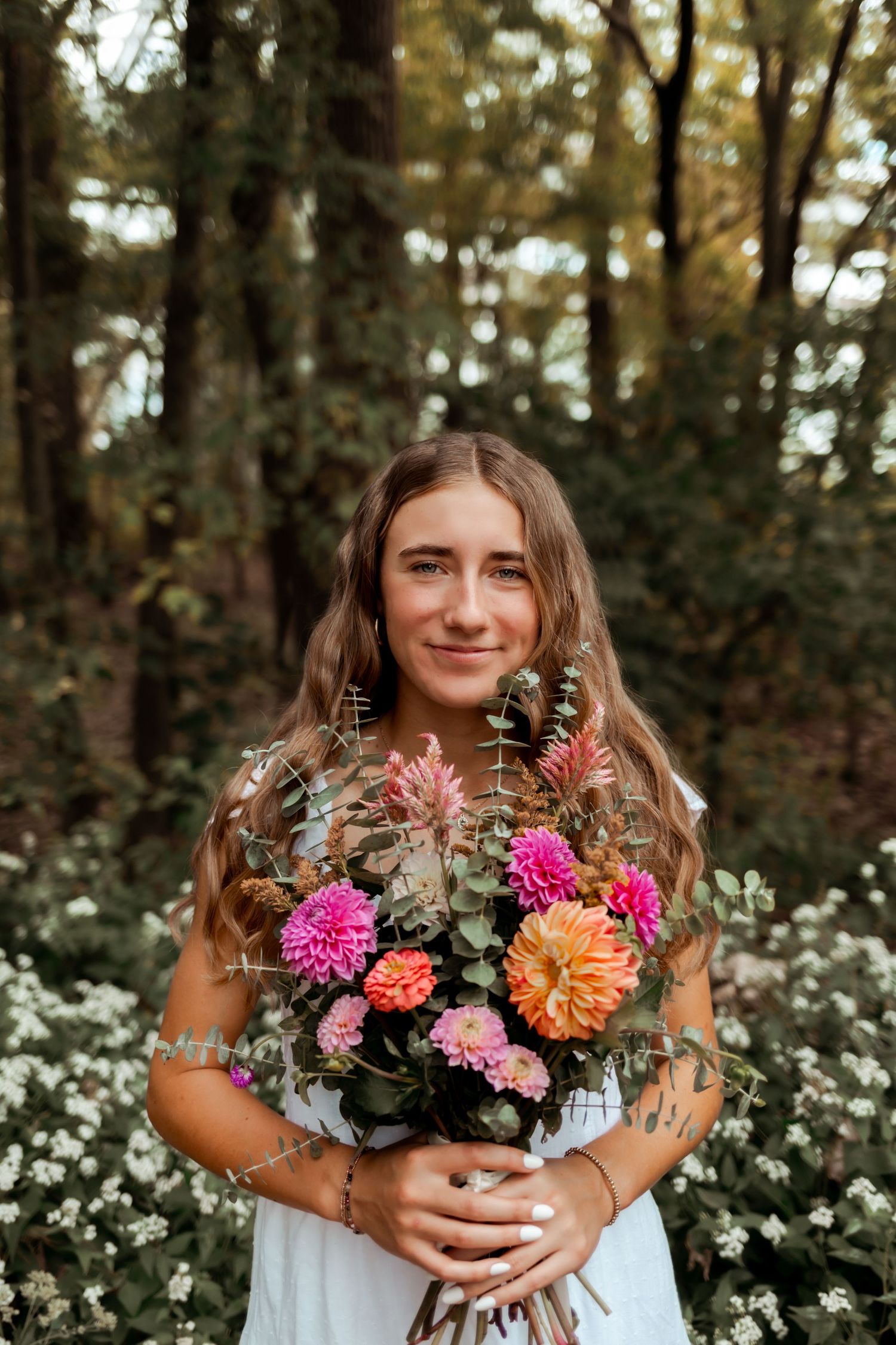 Person holding vibrant bouquet with dahlias and wildflowers in wooded setting.