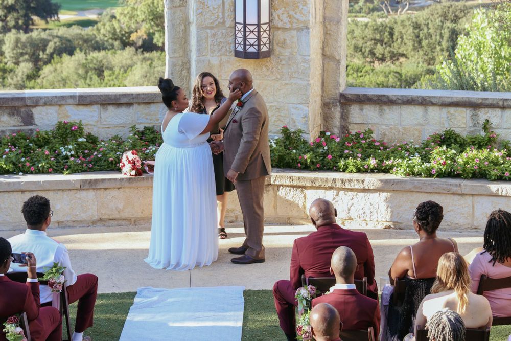 Wedding guests seated on burgundy chairs watch ceremony under stone archway.