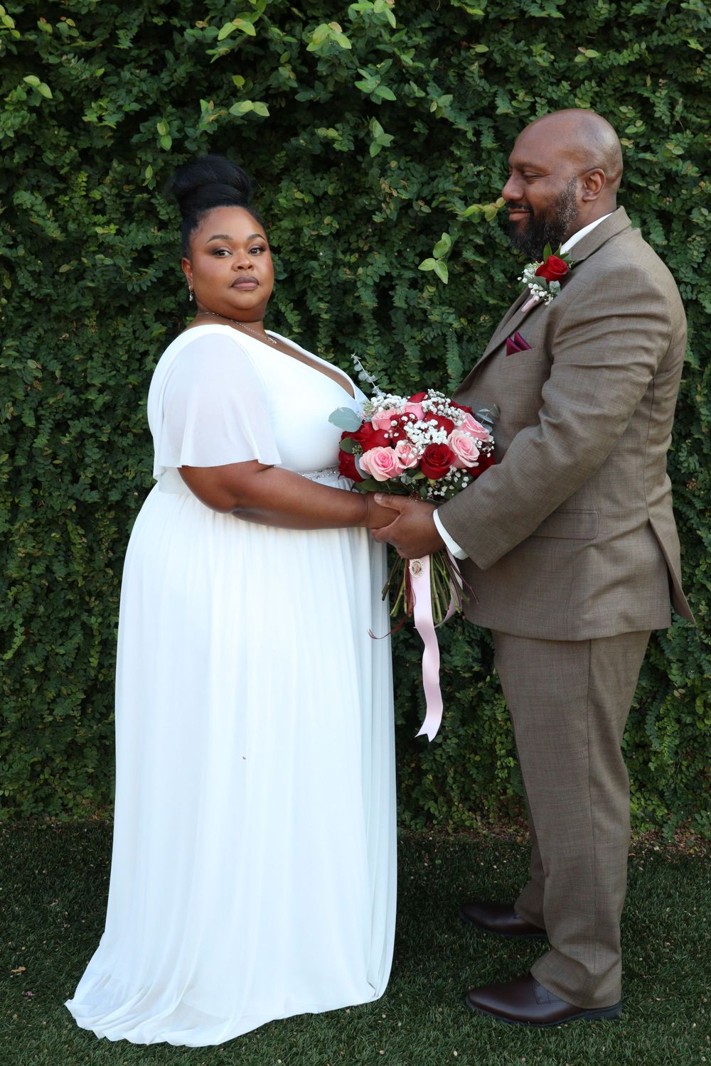 Bride in white dress and groom in tan suit pose with floral bouquet against green hedge.