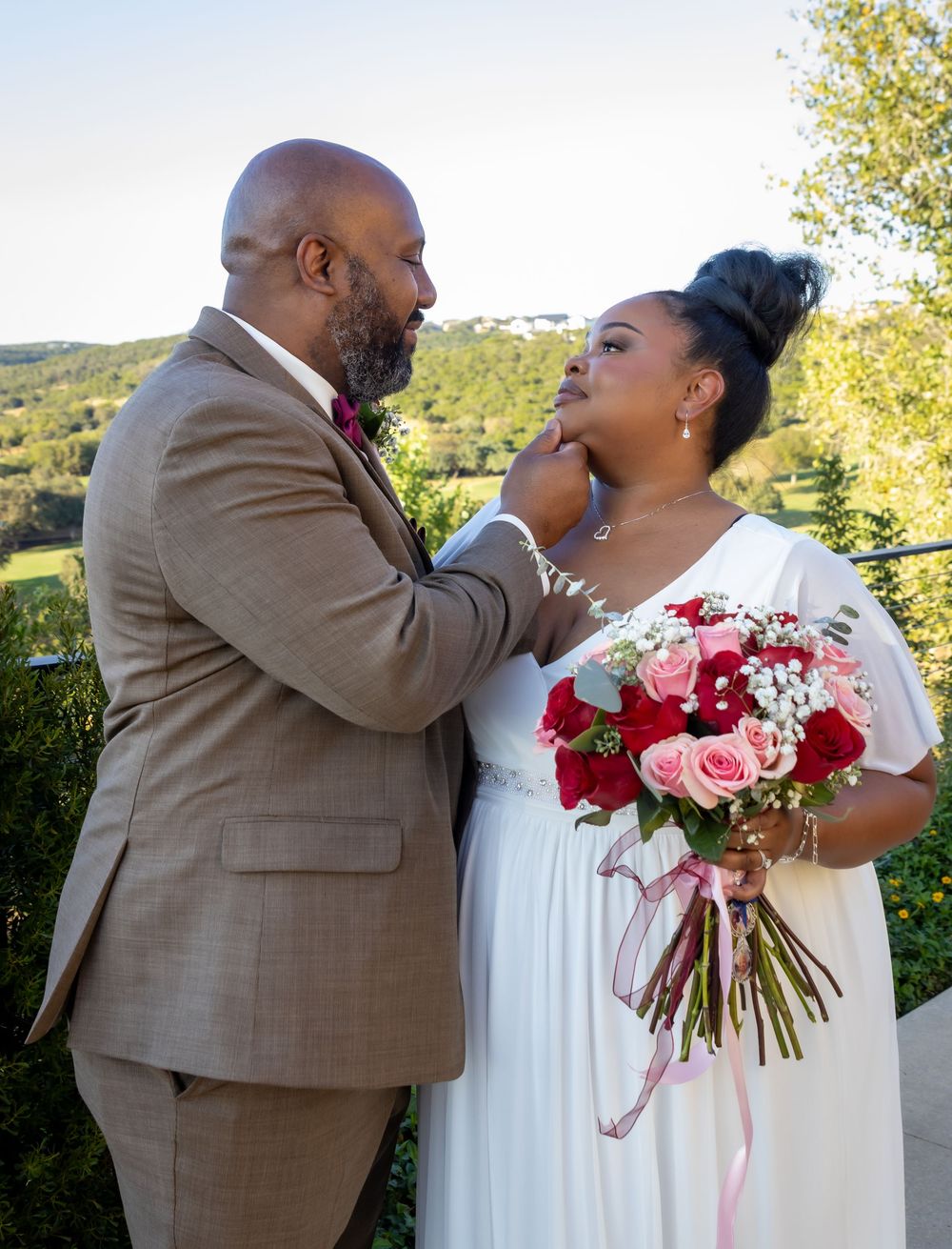 A couple shares a tender moment during their outdoor wedding ceremony with a beautiful pink and red rose bouquet.
