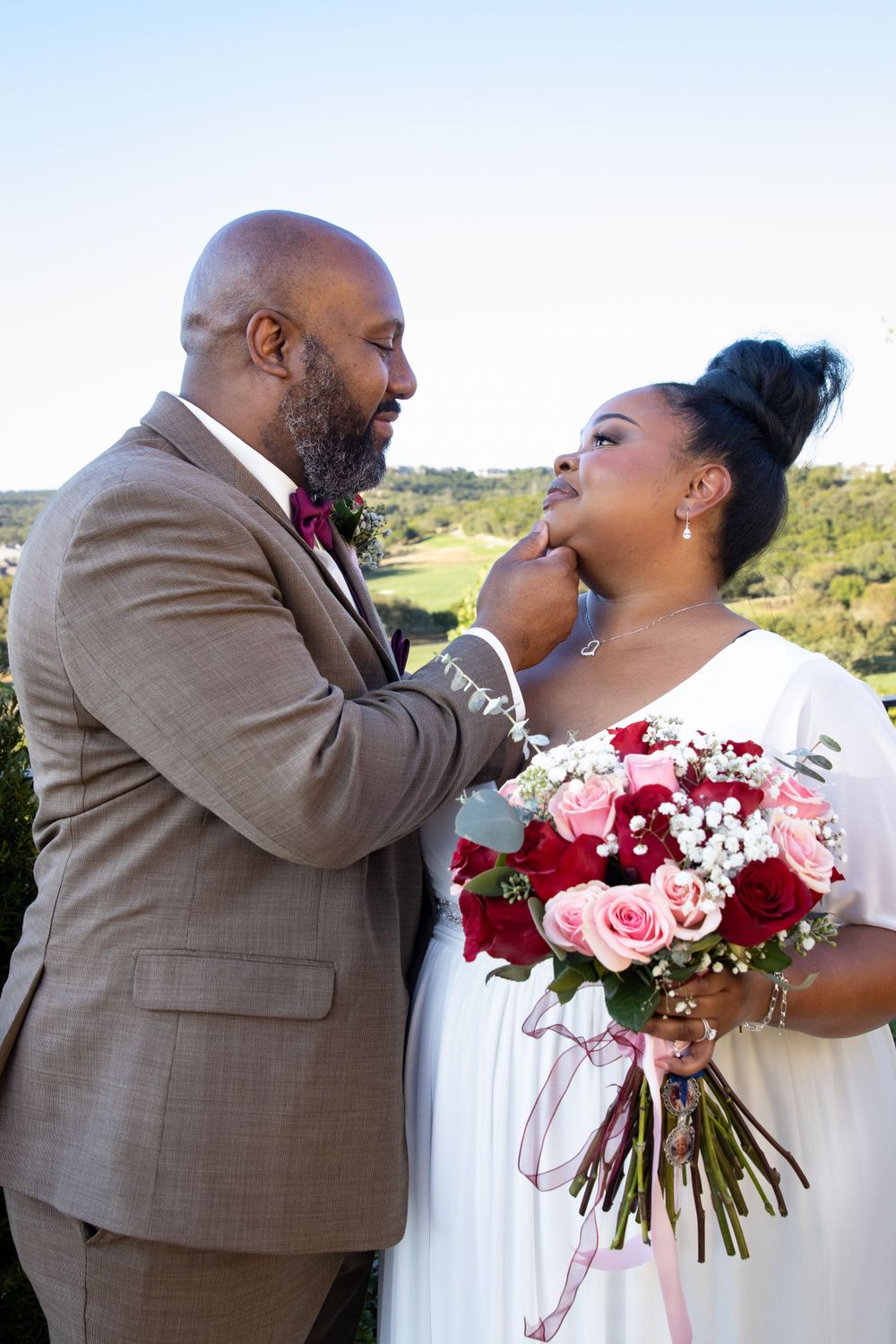 Groom in tan suit tenderly touches bride's face while she holds red and pink bouquet.