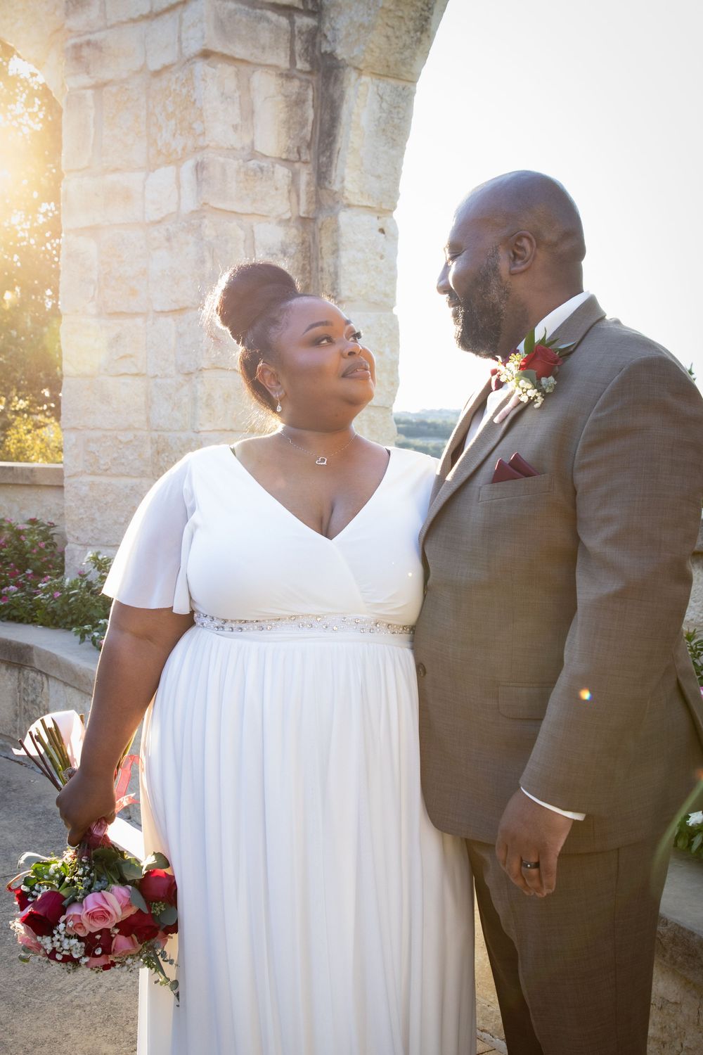 Newlyweds share loving glance in golden sunlight under stone archway.