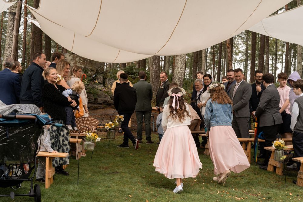 White tent canopy set up in a forest setting with guests seated on wooden benches for an outdoor event.