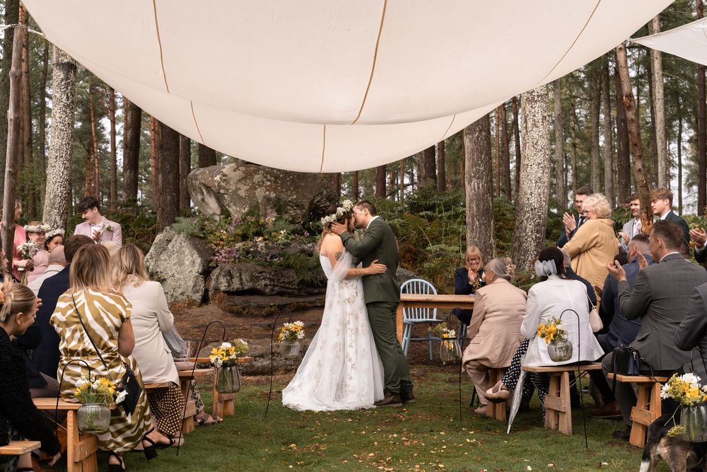 A romantic outdoor wedding ceremony under a white canopy in a forest setting with guests seated on wooden chairs.