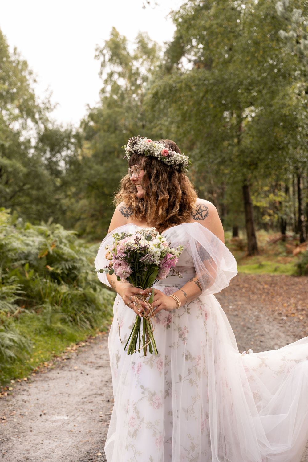 Two people in flowing white dresses hold floral bouquets while walking down a woodland path together.