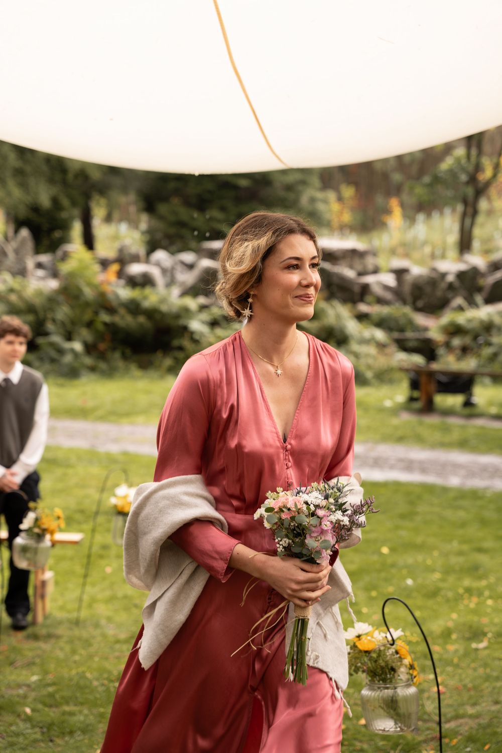 A bridesmaid in a dusty rose dress holds a bouquet while walking outdoors at a garden wedding ceremony.