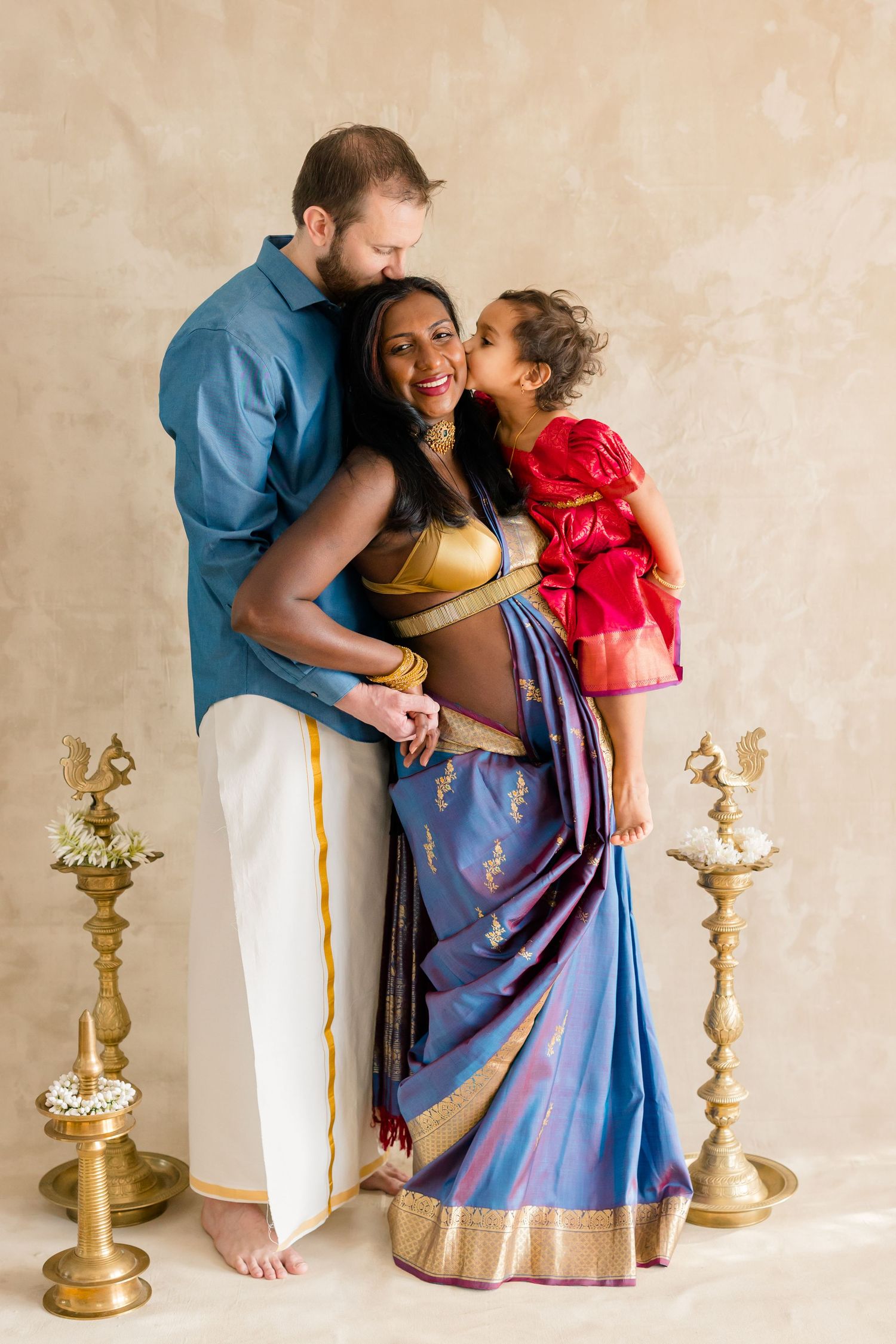 A loving family portrait with traditional Indian dress elements and decorative brass lamps against a neutral backdrop.