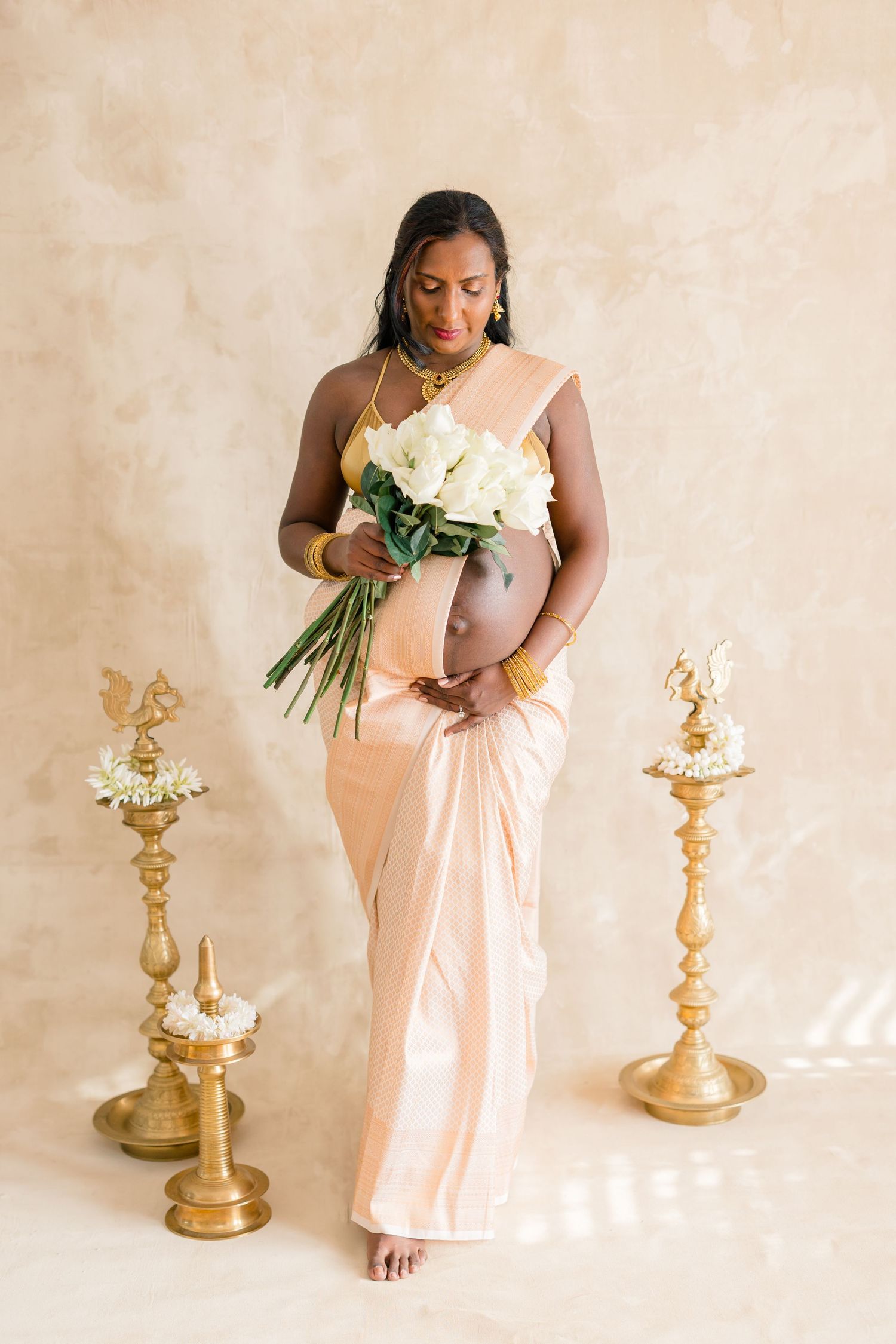 Elegant maternity photo session featuring a soft peach dress, white calla lilies, and gold candlesticks against a neutral backdrop.