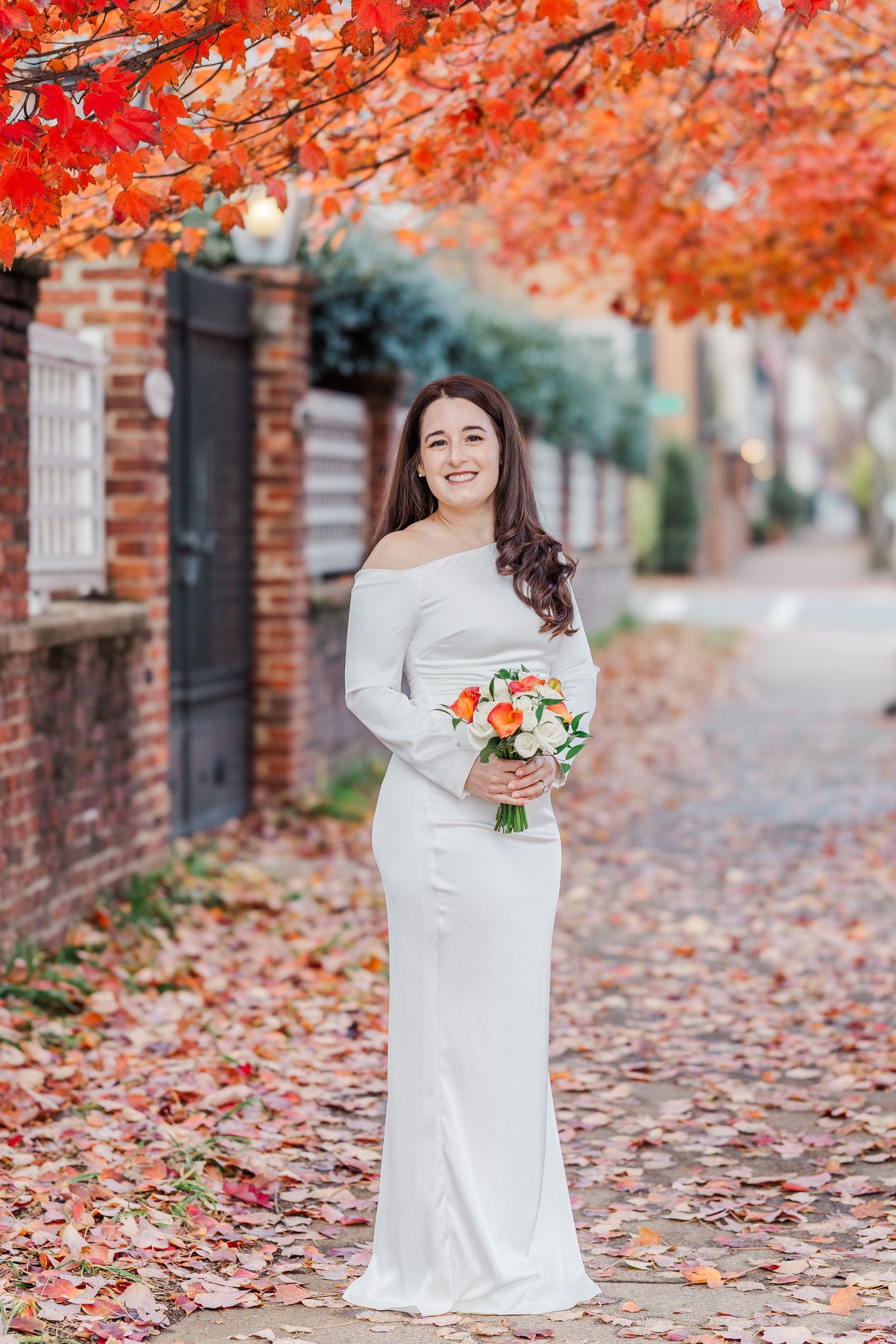 Elegant white wedding gown against vibrant autumn foliage with red maple leaves and brick wall backdrop on city sidewalk.