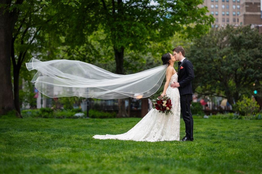 A bride's veil flows dramatically in the wind against a green park backdrop during a wedding at Washington Square Park in Philadelphia
