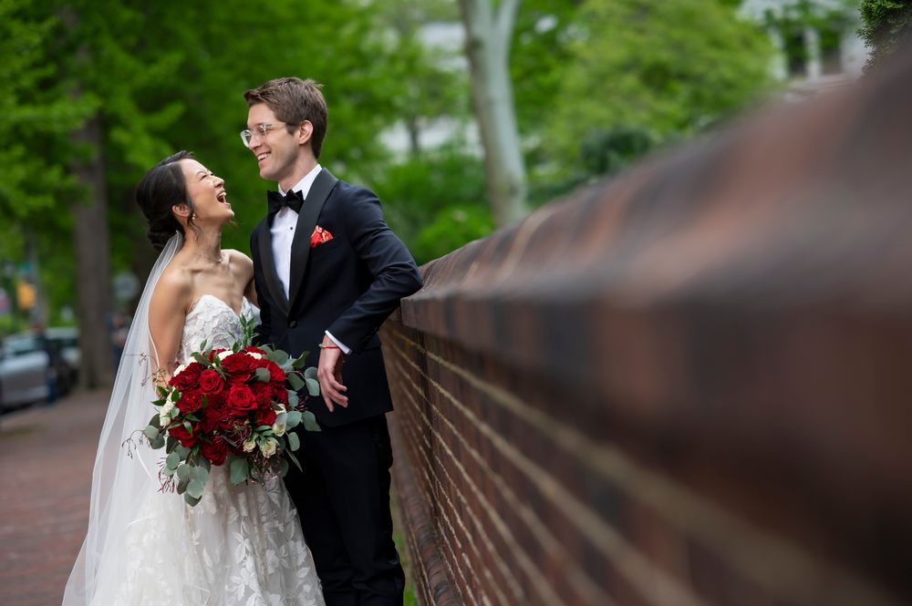 A bride and groom share a romantic moment on a rustic wooden bridge surrounded by lush green trees in Washington Square Park in Philly