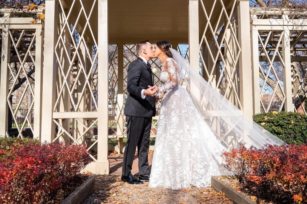 A couple shares a romantic moment under an ornate white lattice gazebo surrounded by red autumn foliage.