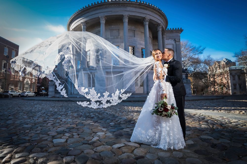A wedding couple poses outdoors in front of a dramatic blue sky next to the Merchants Exchange Building in Philadelphia