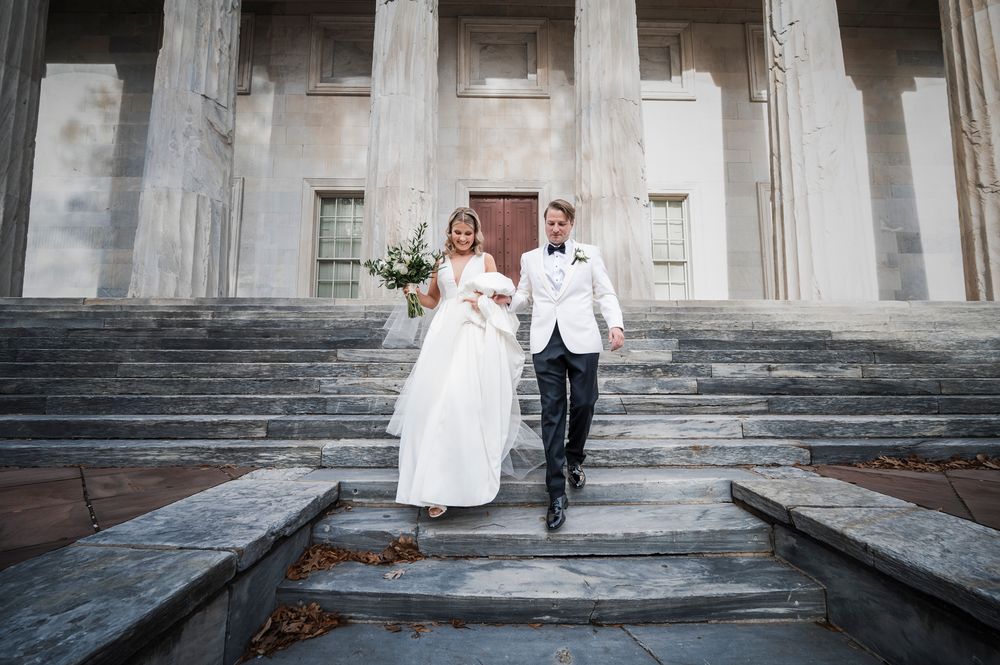 Elegant couple in wedding attire descends stone steps at historic building entrance during their ceremony.